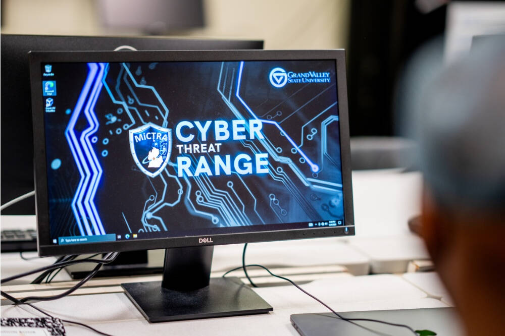 A computer monitor displays a digital interface with the text "CYBER THREAT RANGE" in large letters, along with the logos of MiCTRA and GVSU. The background features a blue and black circuit board design, and the monitor sits on a a white desk.
